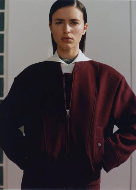 Close-up shot of a woman with dark hair wearing a maroon jacket with a white collar, posing indoors against a light wall.