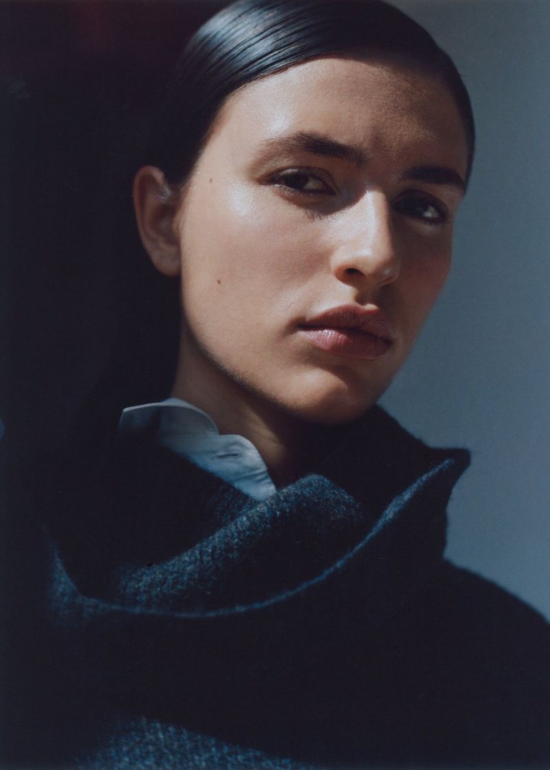 Close-up studio portrait of a fair-skinned woman with dark hair, wearing a dark wool coat, standing against a gray background, under natural light.