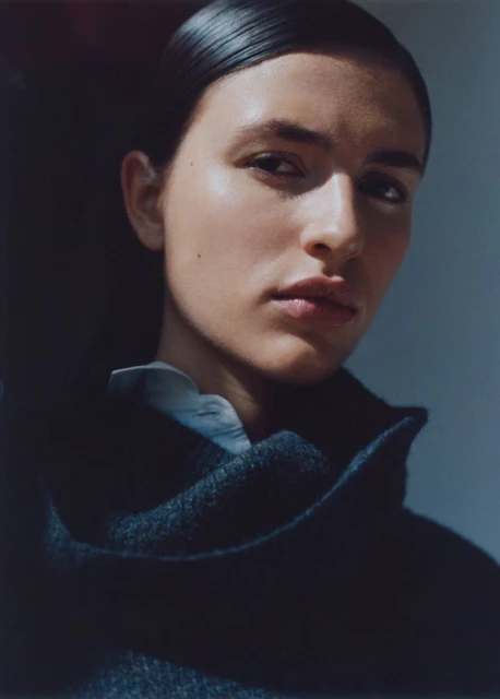 Close-up studio portrait of a fair-skinned woman with dark hair, wearing a dark wool coat, standing against a gray background, under natural light.