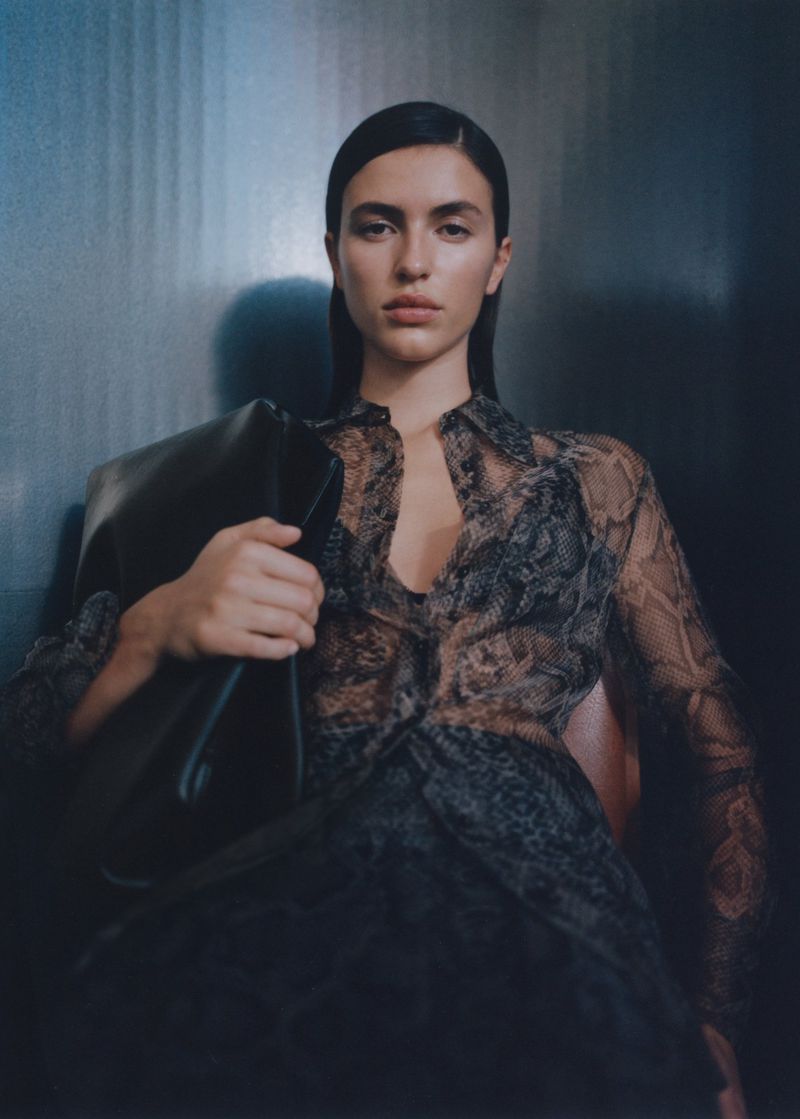 Glamorous woman in sheer black snake-print top holds a large leather handbag, sitting indoors against a textured metal background.