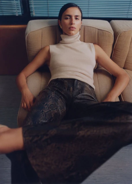 A woman reclines in a beige chair wearing a sleeveless cream turtleneck, snakeskin-print pants, and gold earrings in a relaxed pose.