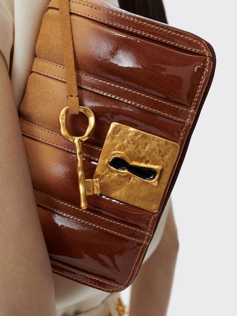 Close-up of a brown leather clutch with gold hardware and a decorative key, against a neutral background.