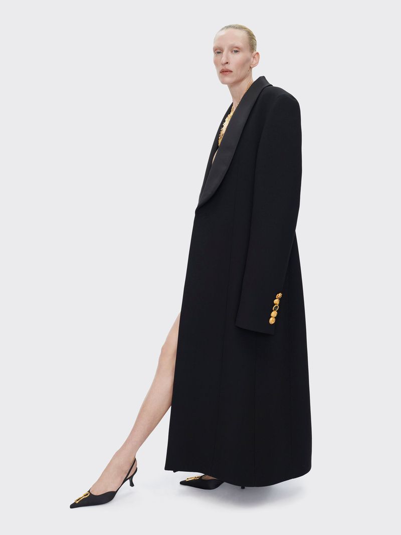 Elegant adult female posing in a long black coat and kitten heels against a neutral backdrop in a studio fashion shot.