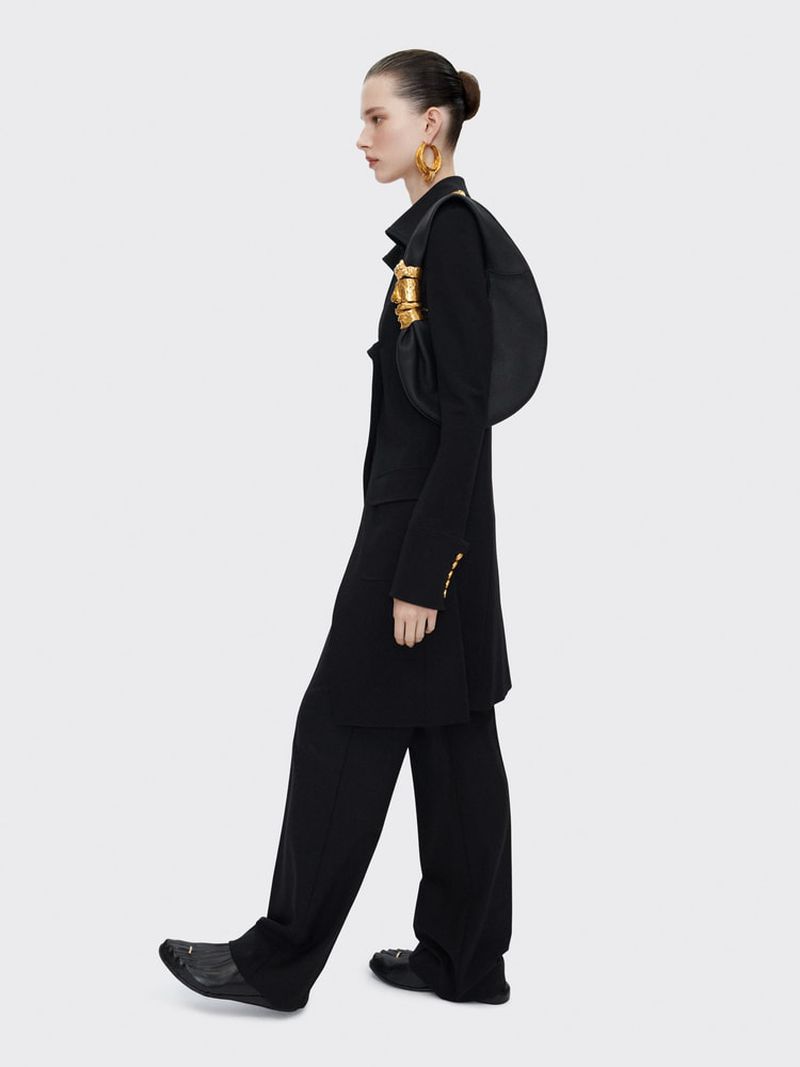 Fashionable adult female strides confidently in an all-black outfit, accentuated by gold earrings and a striking gold detail on her purse.