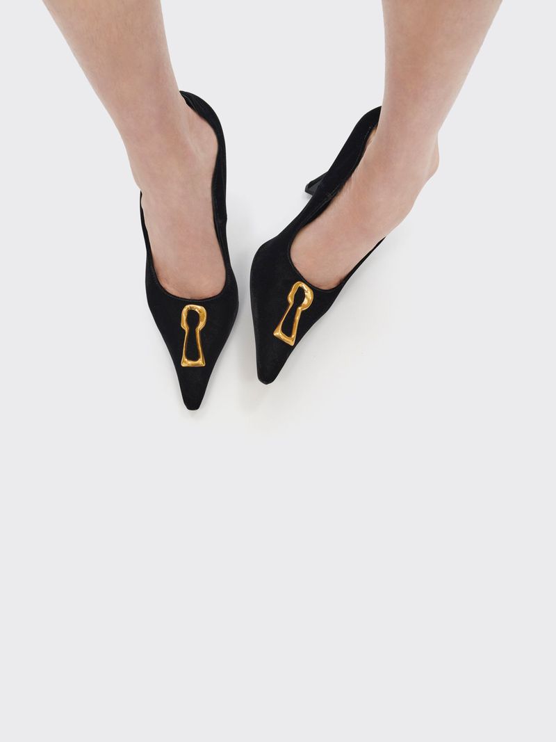Overhead shot of black, pointed-toe heeled mules featuring gold keyhole accents worn by a light-skinned person on a white surface.
