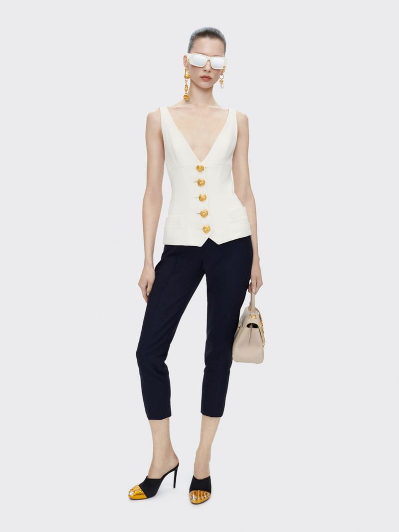 A slender woman poses in a sleeveless cream top with gold buttons, cropped black trousers, sunglasses, gold earrings, and black-gold heels.