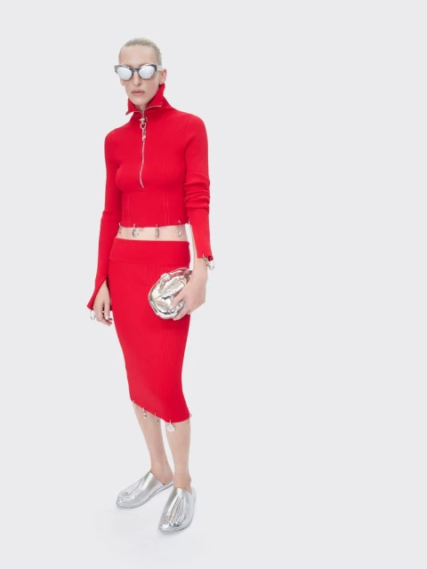 A slender woman poses in a bright red, form-fitting knit top and skirt, accessorized with silver shoes and a silver clutch against a white backdrop.