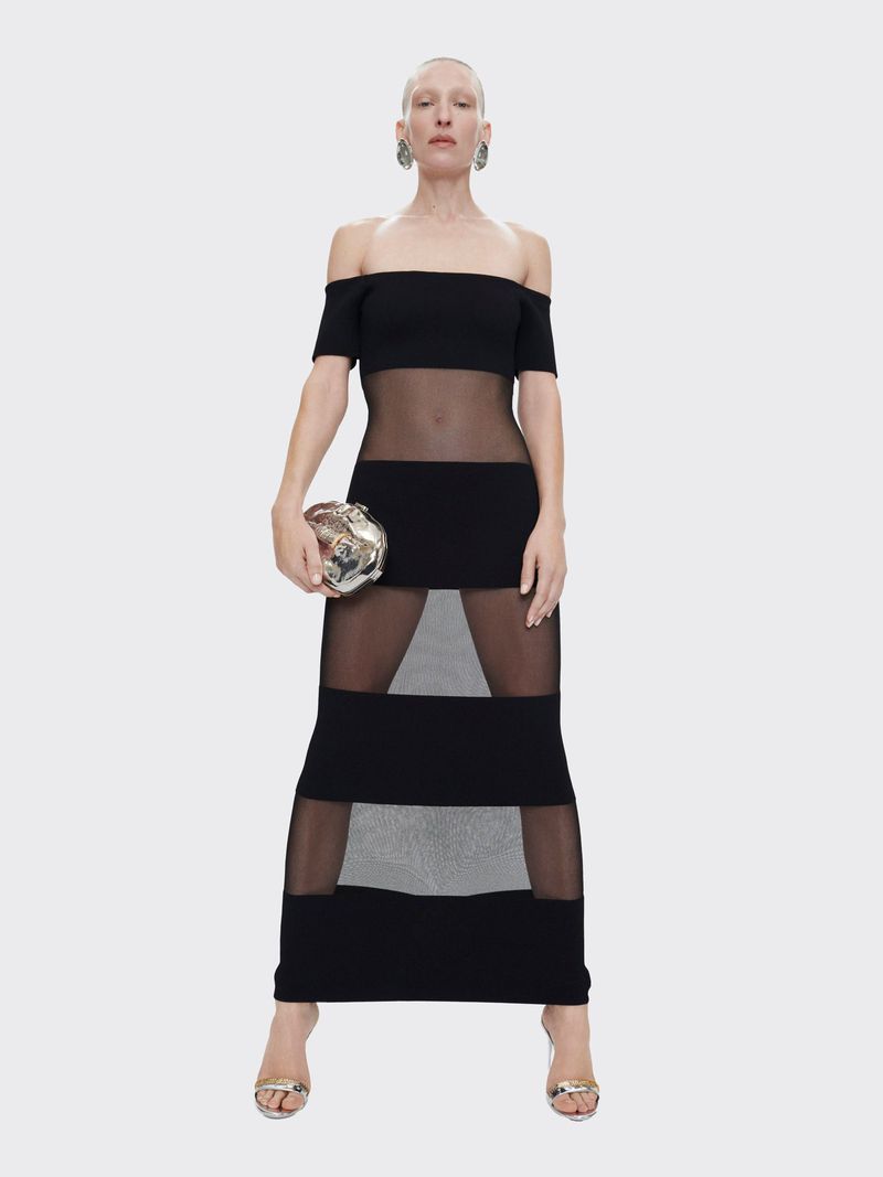 A slender adult female poses in a black off-shoulder dress with sheer mesh panels, accessorized with metallic heels and a silver clutch.