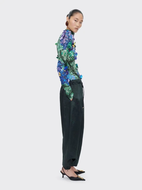 Full-length studio shot of a slender Asian woman wearing a floral top, loose black pants, and black kitten heels against a white background.