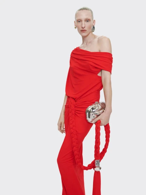 Woman wearing a red gown with a braided rope accent and silver clutch bag, posing in a studio setting with a plain background.
