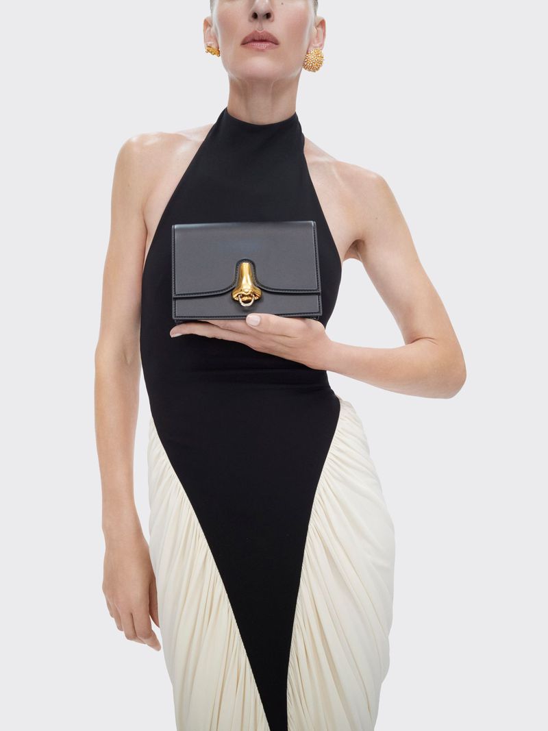 Woman in black and white dress holding a small black purse with gold hardware against a neutral background in a studio setting.