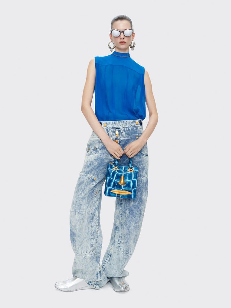 Studio shot of a fashion model posing in a blue top and patterned denim jeans, holding a blue purse with a face, wearing silver shoes.