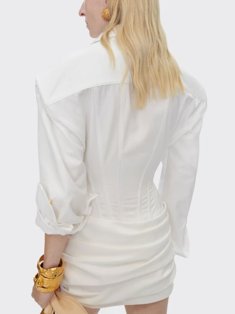 Woman wearing white blouse and skirt, gold bracelet and earring with light skin, shot from behind in front of white background.