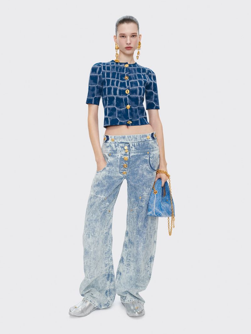 A model poses in a blue patterned top, light wash denim pants, and silver shoes against a white background in a full studio shot.