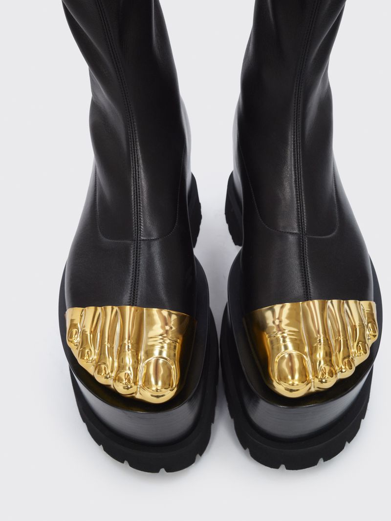 High-angle view of black platform boots with sculpted golden feet adorning the toes against a seamless, light gray backdrop.
