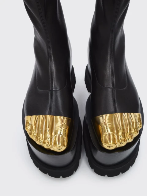 High-angle view of black platform boots with sculpted golden feet adorning the toes against a seamless, light gray backdrop.