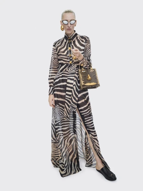 Woman in a zebra-print maxi dress and sunglasses poses in a studio with a small gold purse and black shoes; fashion editorial shot.