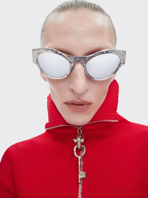 Close-up studio shot of a person with silver reflective sunglasses, red high-collar ribbed shirt, and a decorative key pendant.