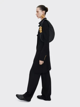 Model in black jacket and trousers wearing statement gold earrings and carrying a black shoulder bag against a white seamless background