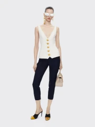 Model wearing a white v-neck vest and dark trousers holding a handbag against a seamless light grey background