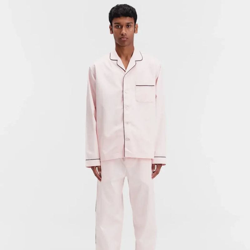 Adult male stands in a light pink, long-sleeved pajama set with dark piping, presented against a plain white backdrop in a studio setting.