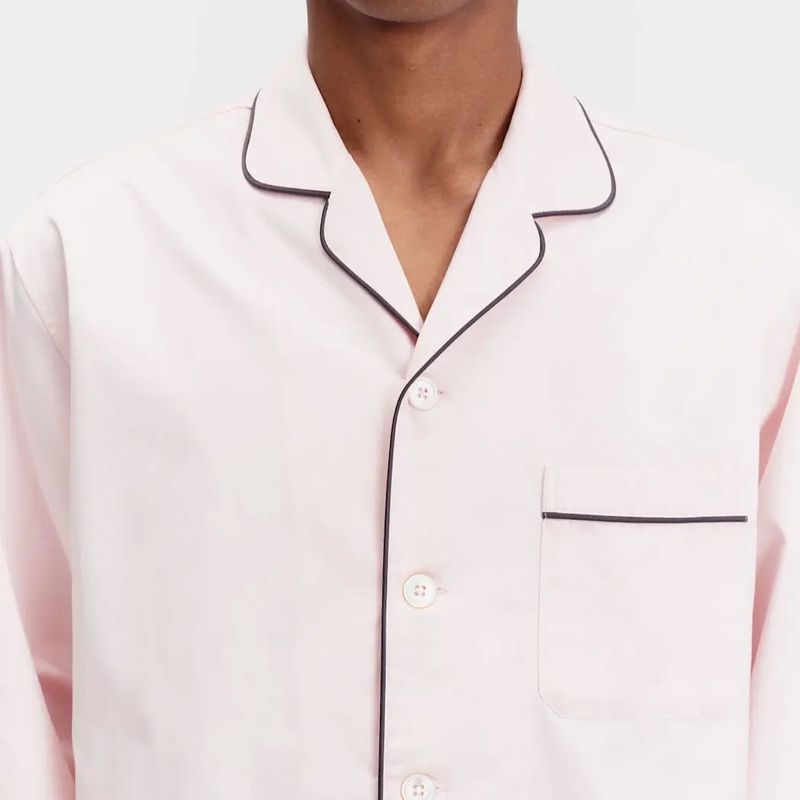 Close-up of a person wearing a light pink, long-sleeved button-down shirt with dark trim on the collar and pocket, in a studio.