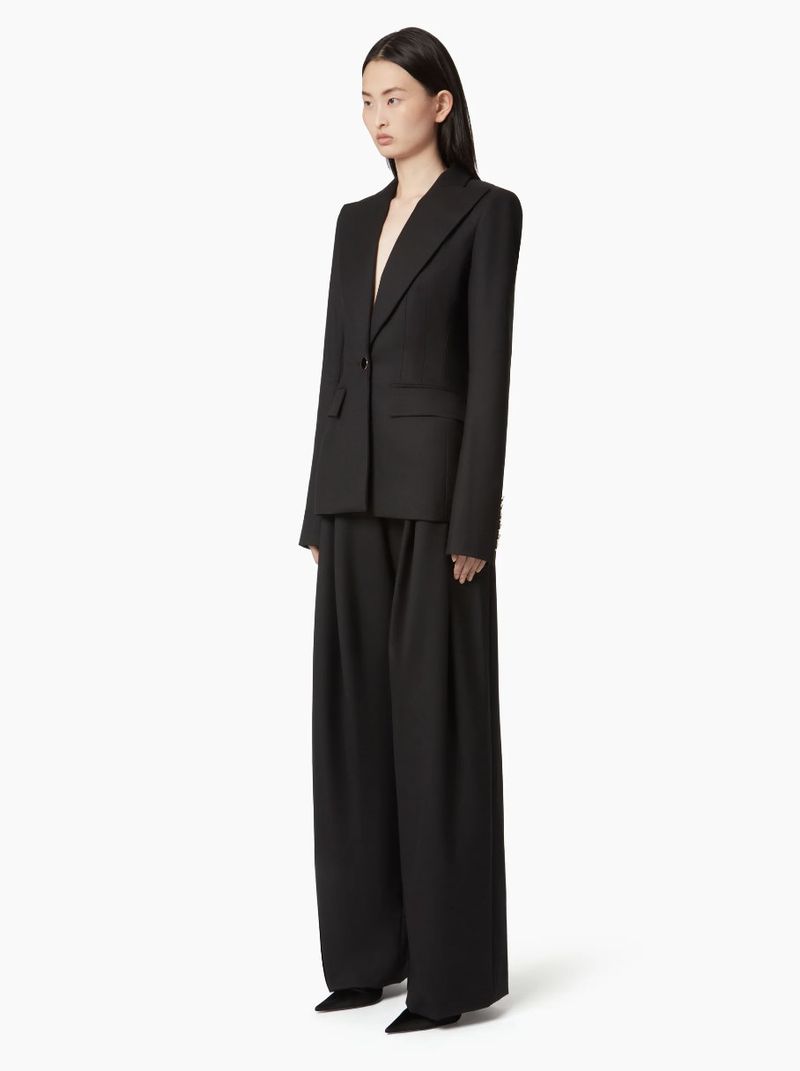 Elegant Asian woman in a black suit with a blazer and wide-leg trousers, standing against a clean white backdrop in a fashion pose.