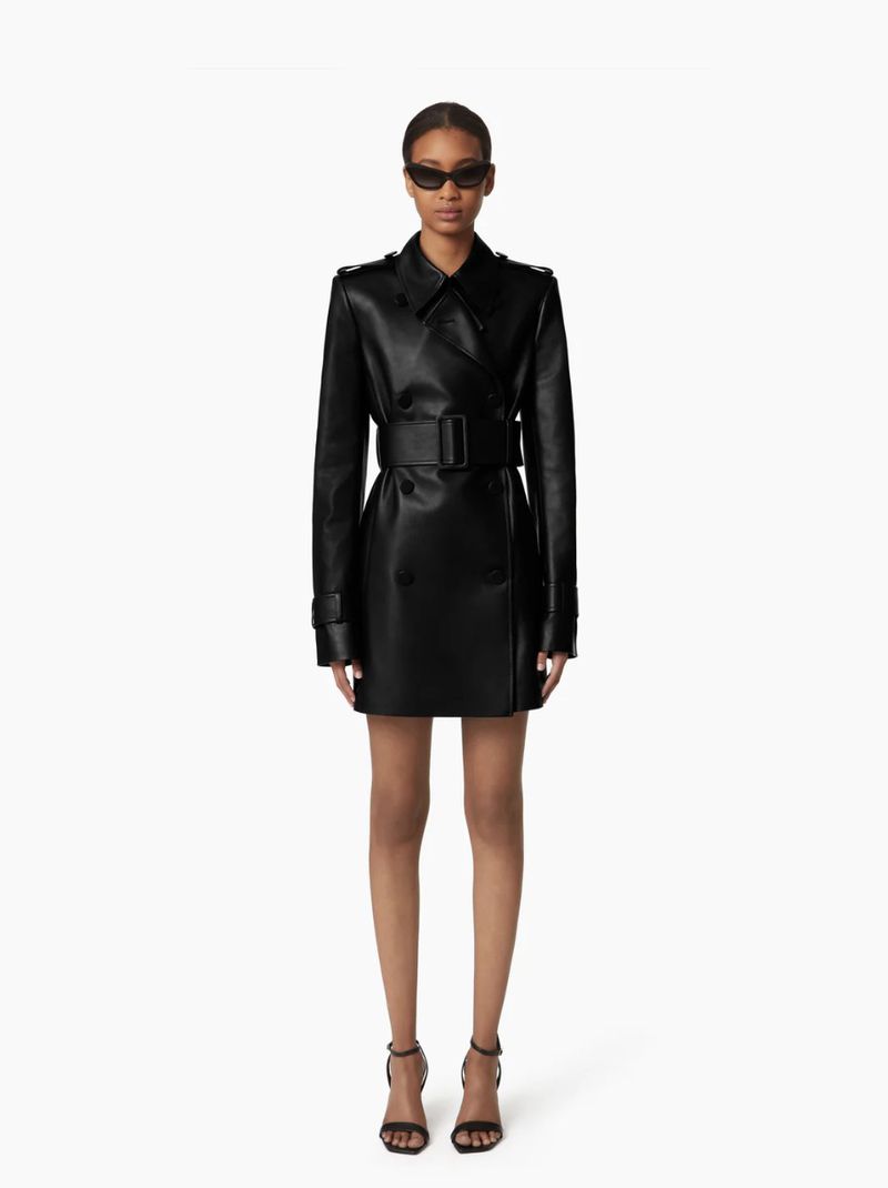 A slender woman poses in a black, double-breasted, belted, knee-length leather coat dress and black strappy heels against a white backdrop.