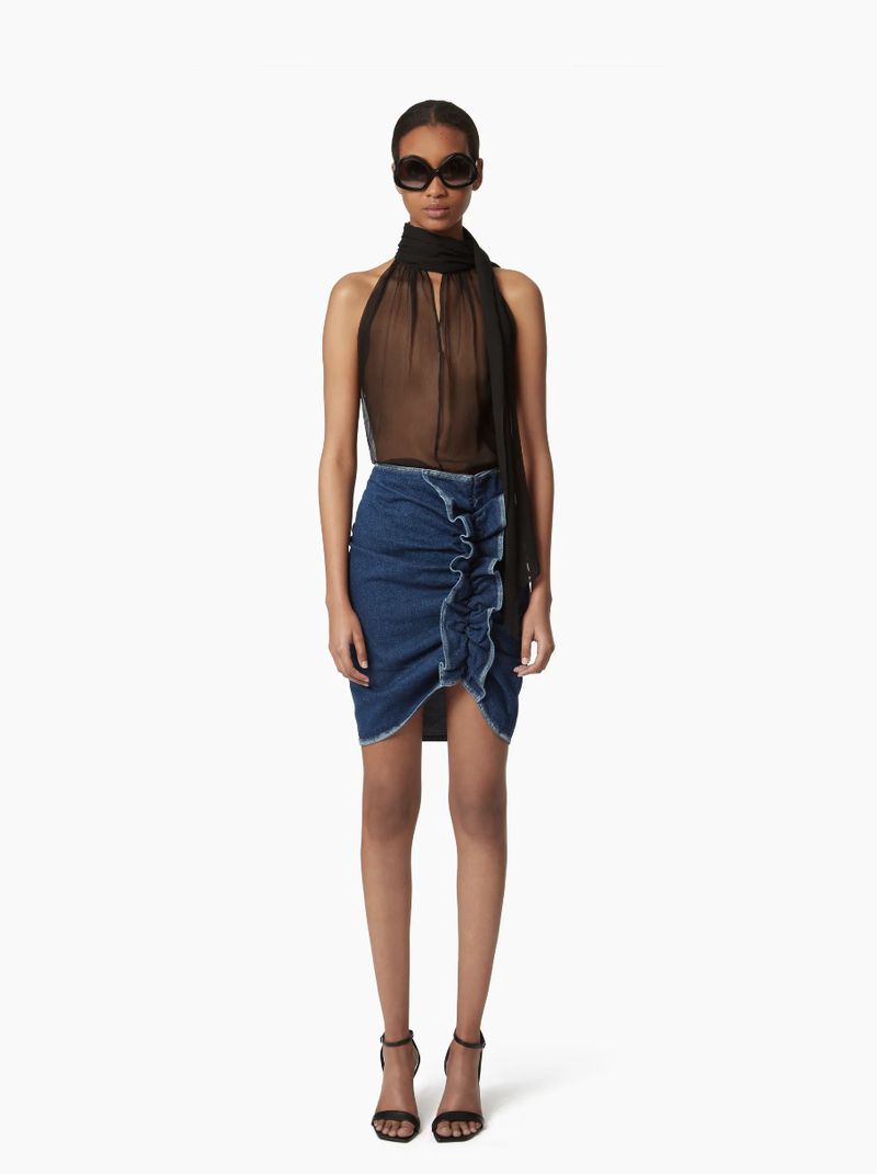 Woman in semi-sheer black halter top, denim mini skirt with ruffle, sunglasses, and heeled sandals stands in a white photo studio.