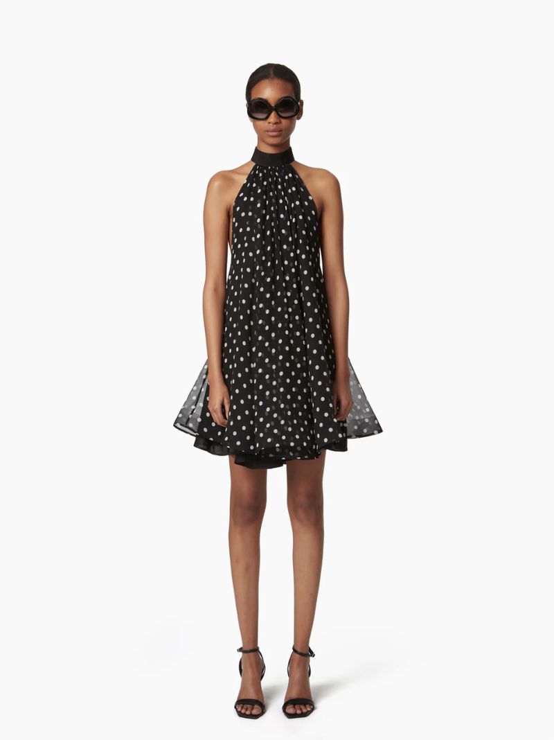 Elegant Black Halter Dress: Woman in a knee-length, polka dot halter dress with sunglasses and black heels stands on a white background.