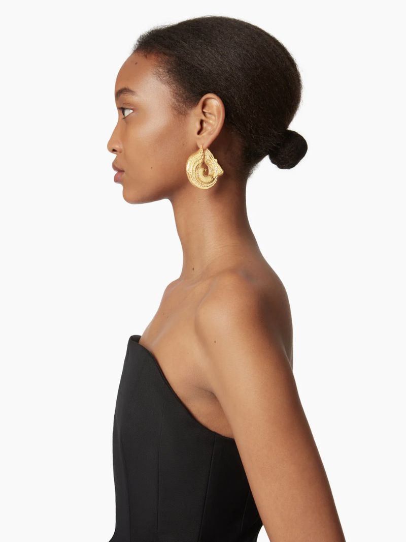 Profile of a woman with dark hair in a bun, wearing a strapless black dress and large gold seashell earrings on a white background.