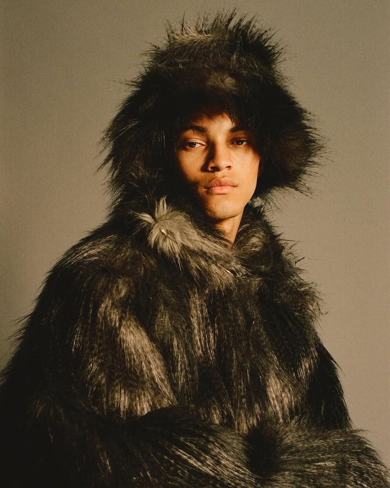 Portrait of a young adult male in a furry hooded coat, posing indoors against a neutral backdrop, captured in soft, diffused lighting.