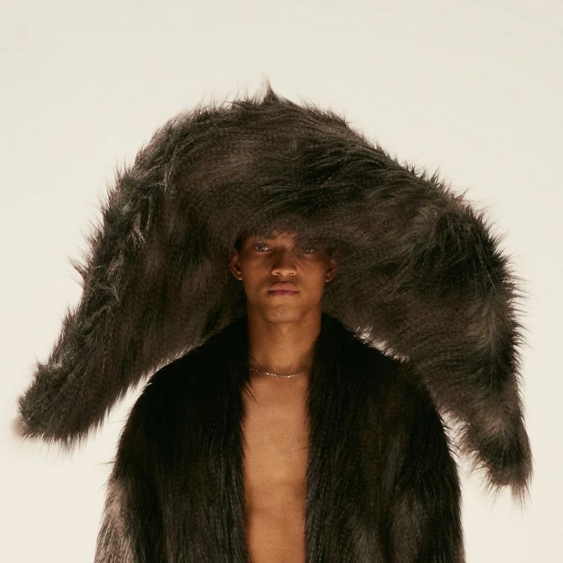 Portrait of a young adult male wearing a shaggy grey fur coat and a giant, oversized fur hat against a plain off-white backdrop.