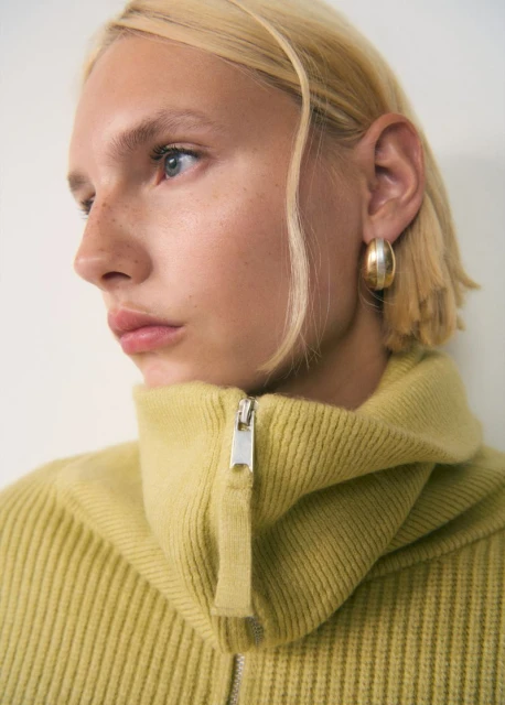 Close-up of a fair-skinned woman with blonde hair wearing a zip-up ribbed sweater and gold hoop earrings against a neutral background.
