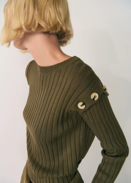 Blonde woman wearing an olive green, ribbed long-sleeve knit sweater with decorative buttons, posing against a white backdrop.