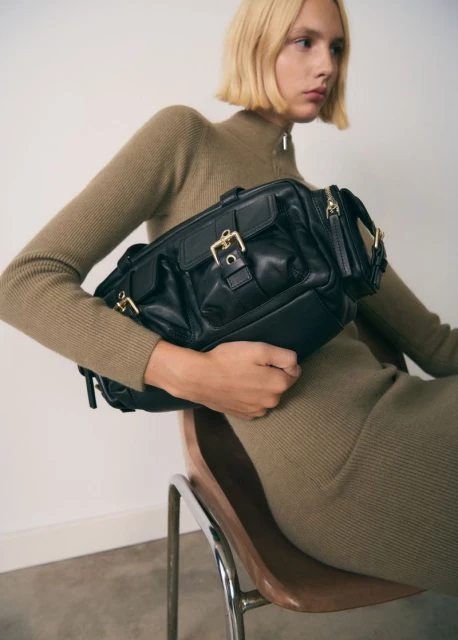 A blonde adult female sits on a chair holding a black leather purse with gold buckles in a studio with neutral background.