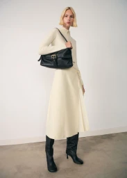 Model in cream knit top and skirt wearing black boots and holding a black leather shoulder bag against a light studio wall.