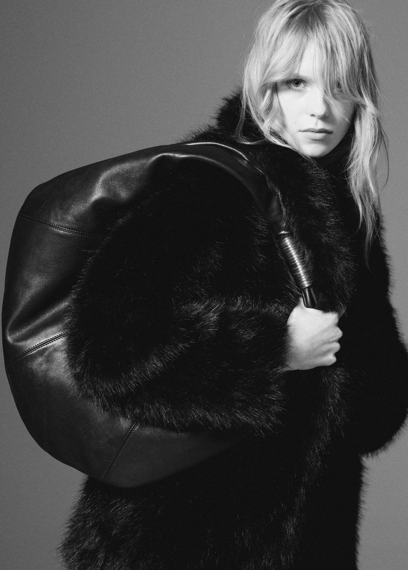 Monochrome studio shot of a blonde woman in a fur coat, carrying a large black leather duffel bag over her shoulder.