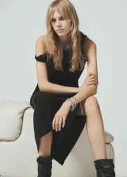 Model wearing black dress and boots seated on white chair against a plain studio backdrop