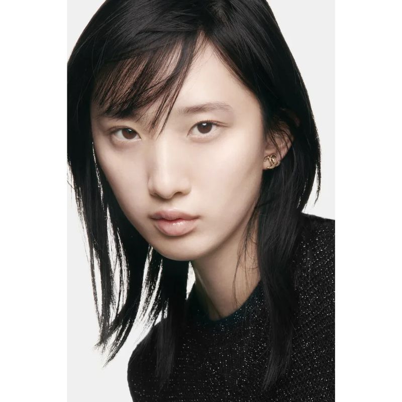 Close-up beauty shot of a young Asian woman with black hair and light skin, wearing subtle gold earrings and a dark textured top.