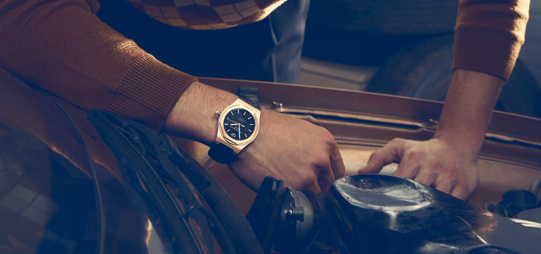 Close-up of a person wearing a gold wristwatch while working on the engine of a car in an indoor environment with cool lighting.