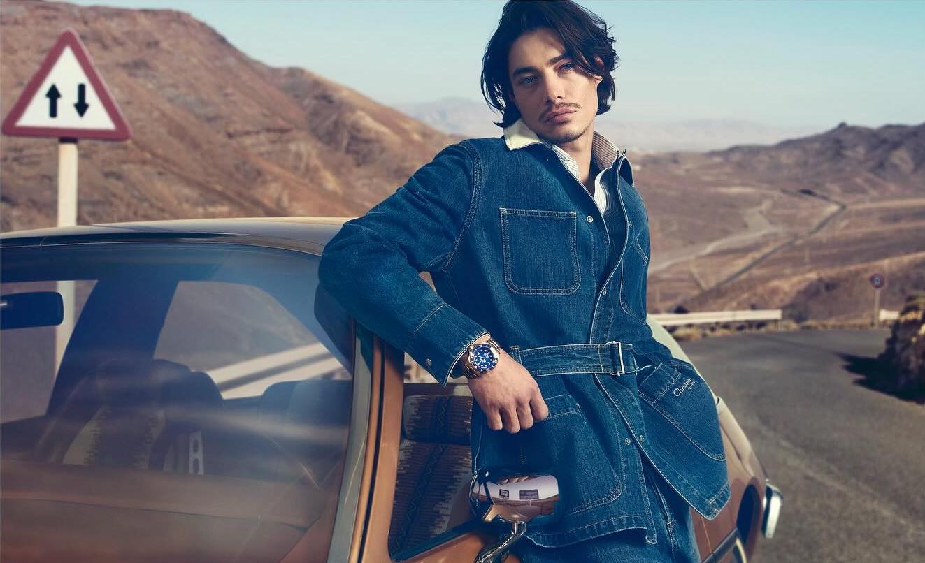An adult male model in a denim jacket leans on a vintage car on a desert road under clear skies, with mountains in the background.