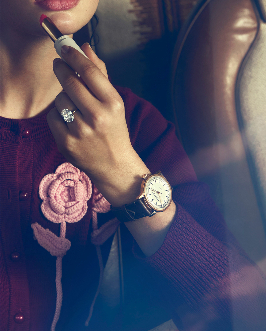 Close-up shot of a woman applying lipstick, wearing a cardigan with a crocheted flower, a watch, and a diamond ring indoors.