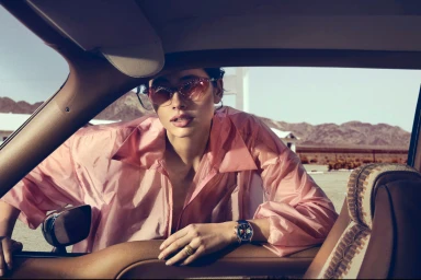 Model leaning into a car window wearing a pink shiny blouse, sunglasses, and a watch against a desert background.