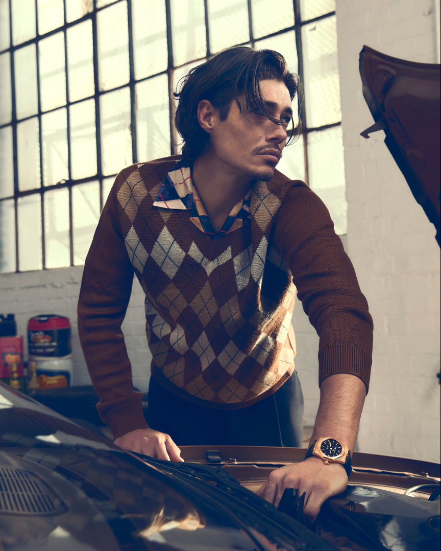 Brunette man in argyle sweater leans over classic car engine in mechanic-style garage, wearing gold watch, soft natural light.