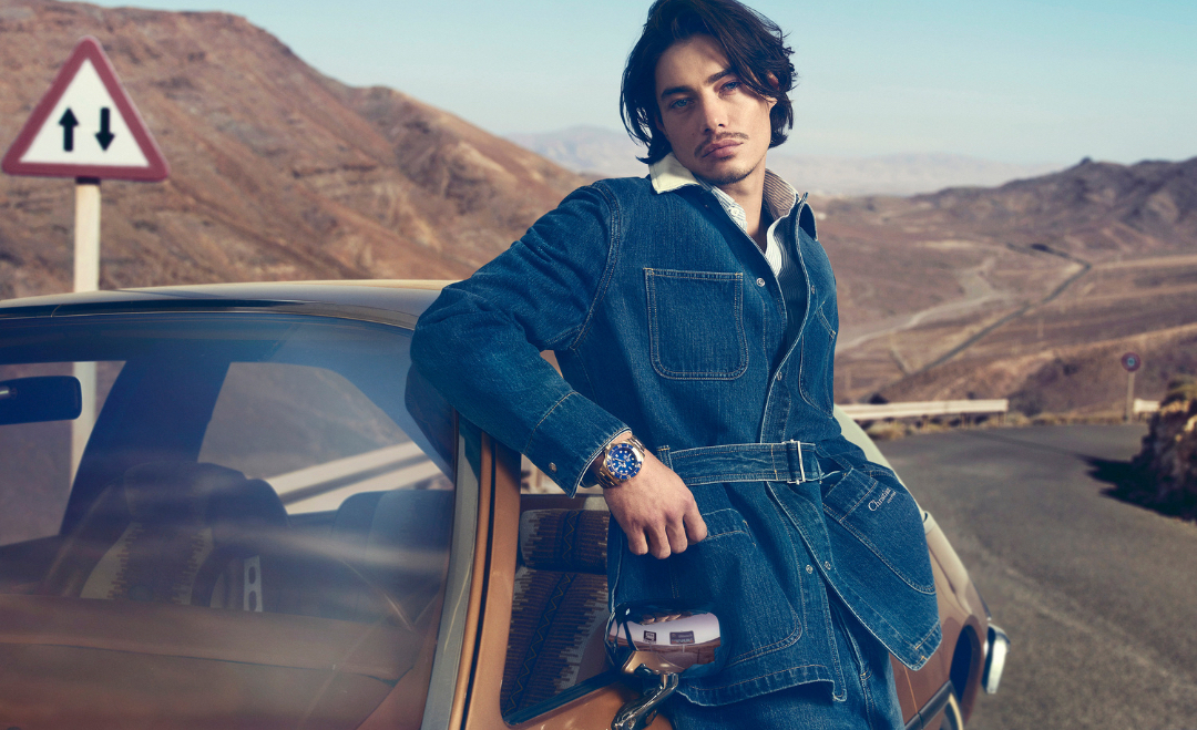 Portrait of a handsome adult male model in denim jacket leaning on a car in a desert setting with a road and distant mountains.