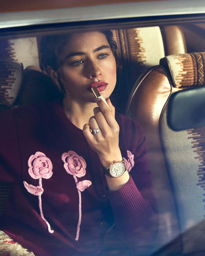 A brunette woman applies lipstick inside a vintage car with patterned seats, wearing a burgundy cardigan with floral appliques and a wristwatch.