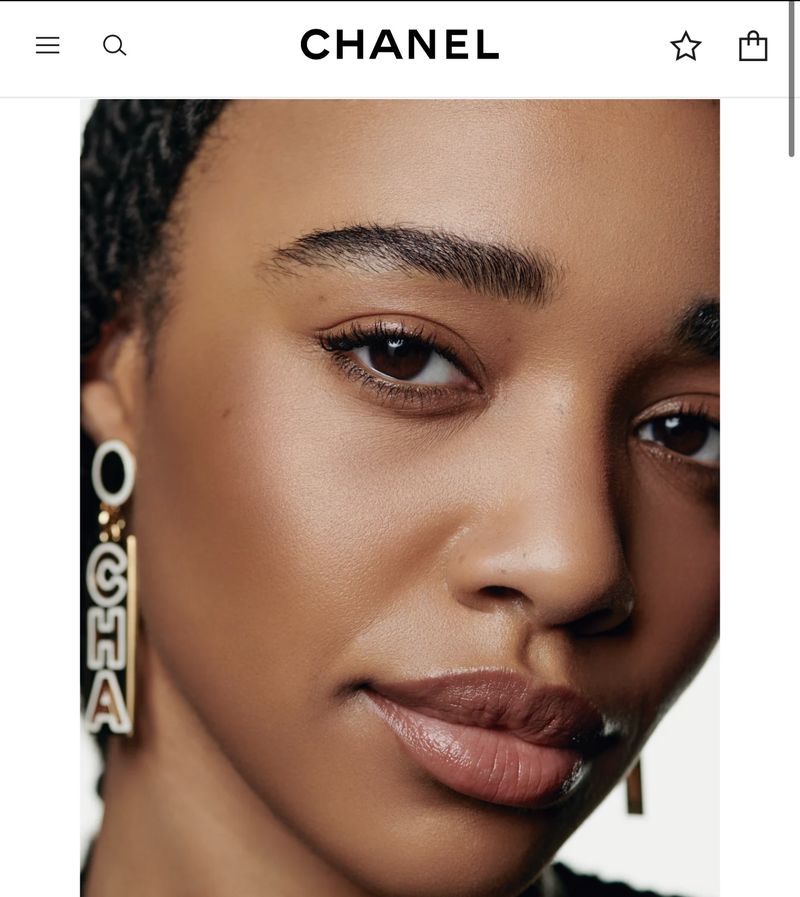 Close-up studio portrait of a dark-skinned woman with glossy lips, wearing statement earrings with CHANEL text, and neutral makeup.