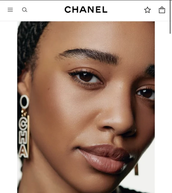 Close-up studio portrait of a dark-skinned woman with glossy lips, wearing statement earrings with CHANEL text, and neutral makeup.