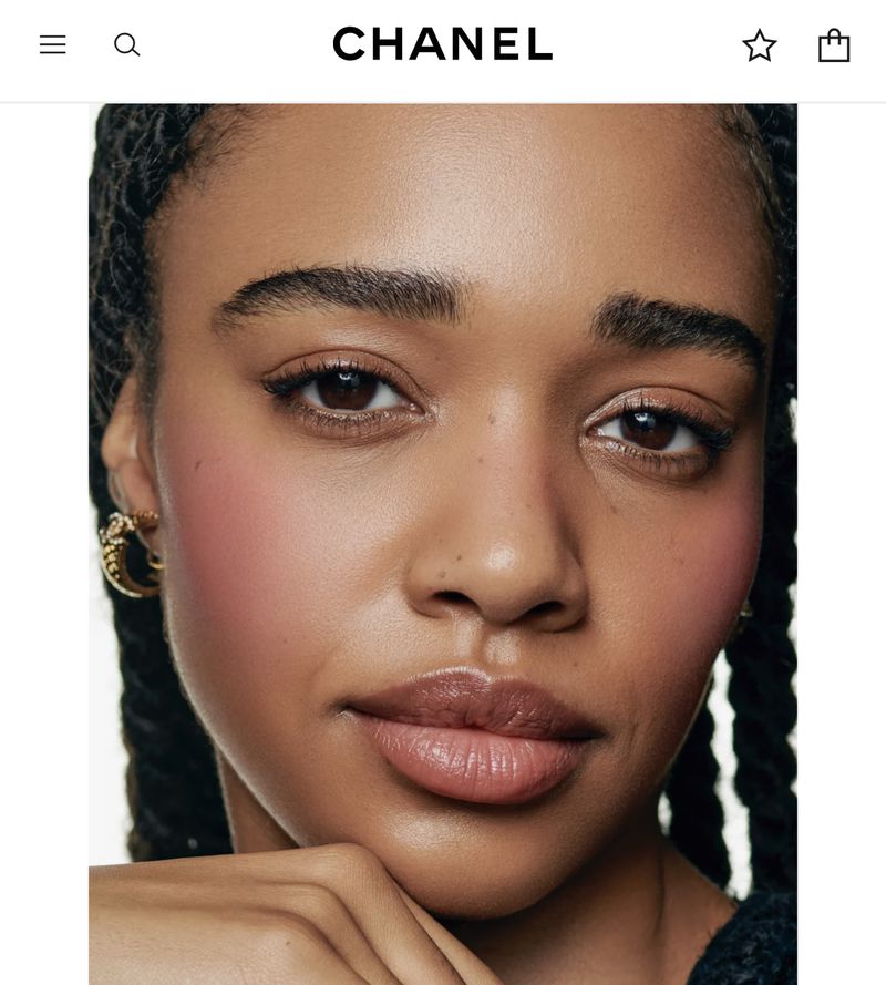 Close-up portrait of a Black woman with light pink blush, gold earrings, and braided hair against a white background with the Chanel logo.